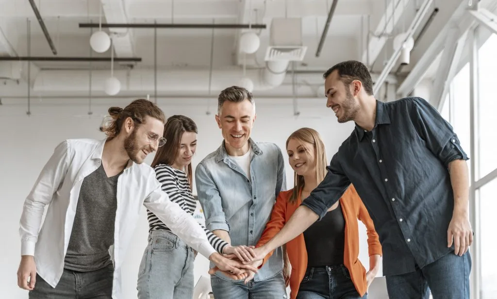 Five office workers, three men and two women, stand together with their hands stacked in the center, celebrating teamwork with smiles and enthusiasm.