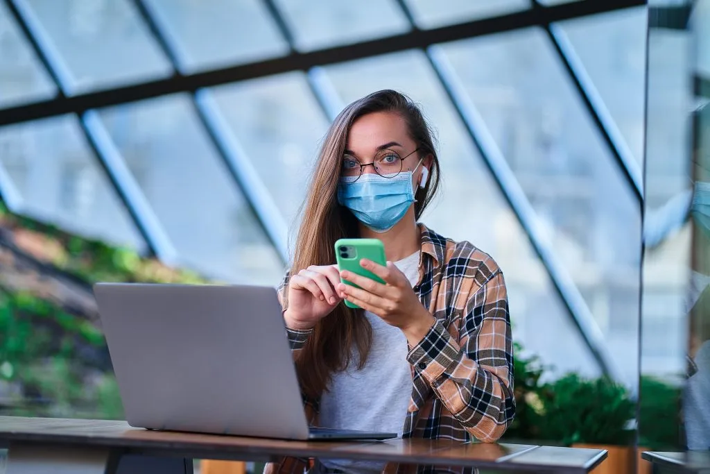 Girl wearing a mask, holding a phone, sitting and working with a notebook.