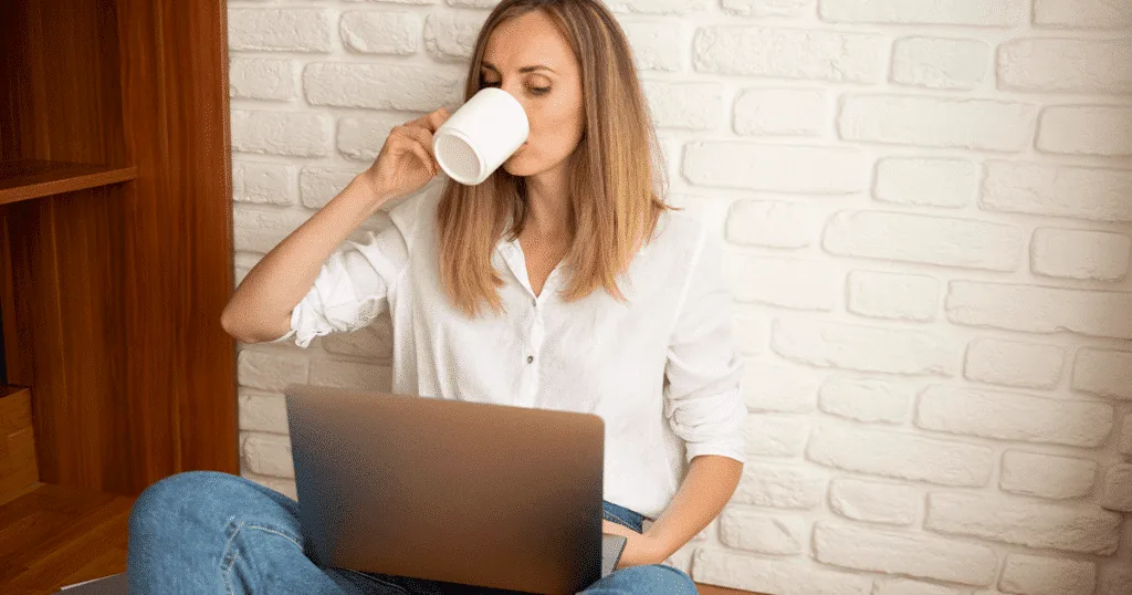 Young woman sitting on the floor, drinking a coffee and working on a laptop, with a relaxed and focused expression.