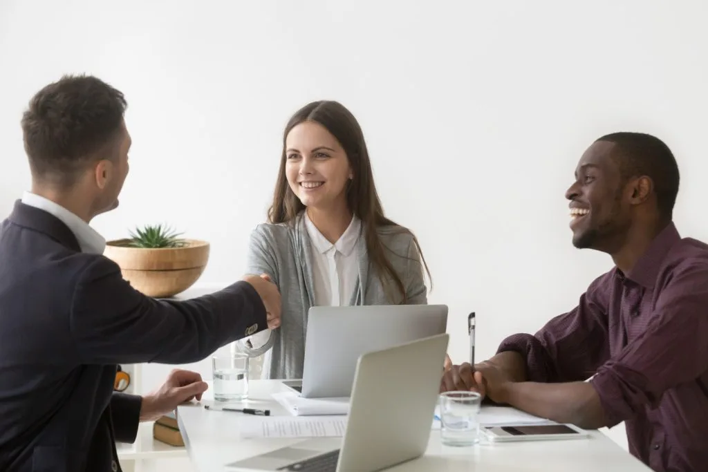 Smiling businesswoman with two male colleagues, one shaking hands and the other laughing, in an office setting.