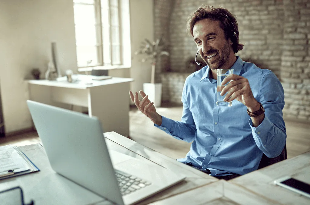 Man smiling at the computer screen while lifting a glass of water