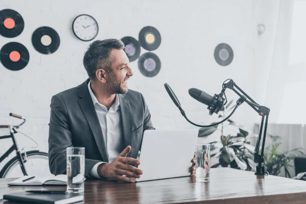 A man is sitting behind a desk with an open notebook in front of him, speaking into a microphone positioned nearby.